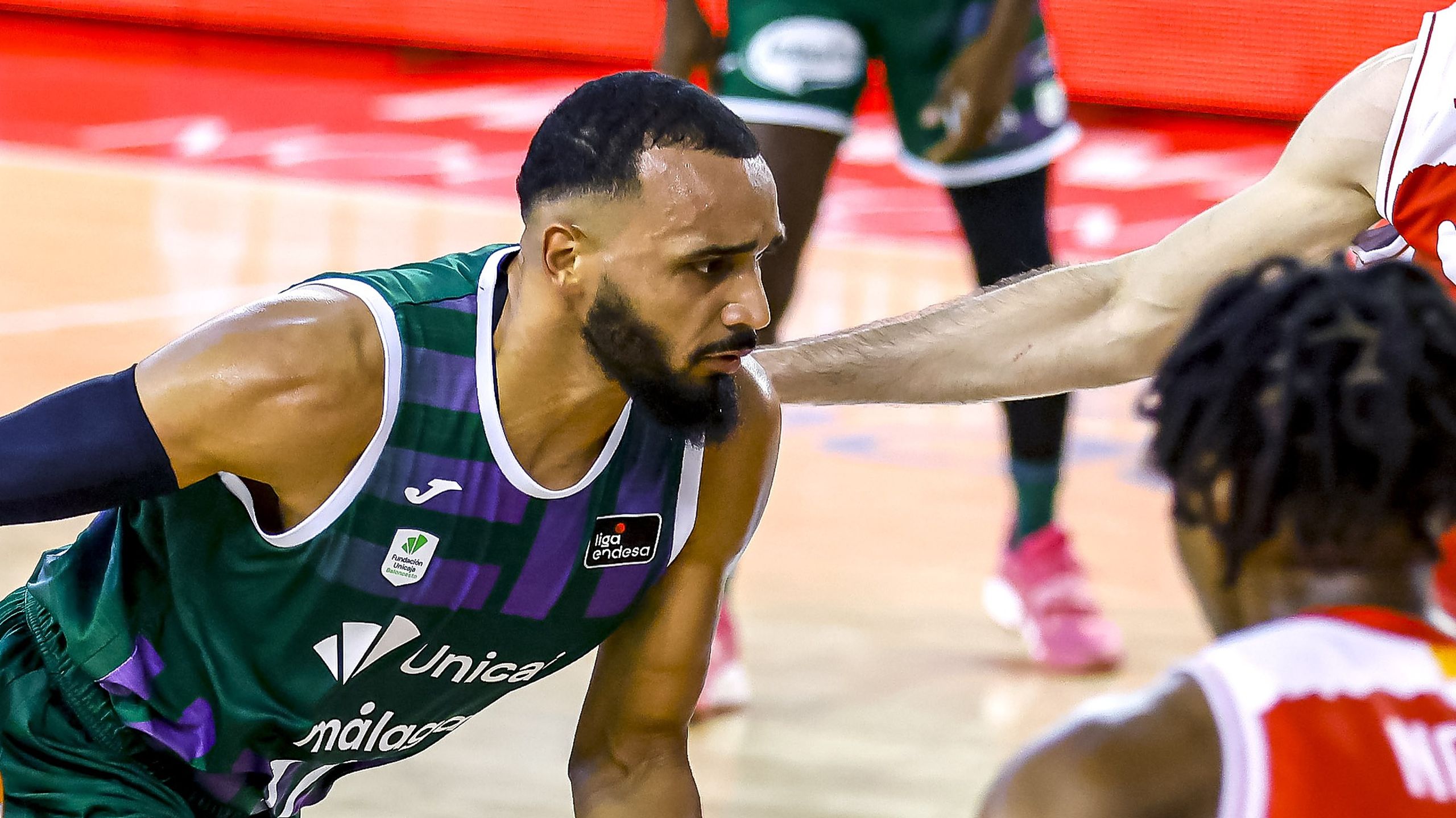 Amine Noua, con la camiseta verde y morada del Unicaja, inclinado hacia delante mientras un rival de camiseta roja extiende el brazo ante él