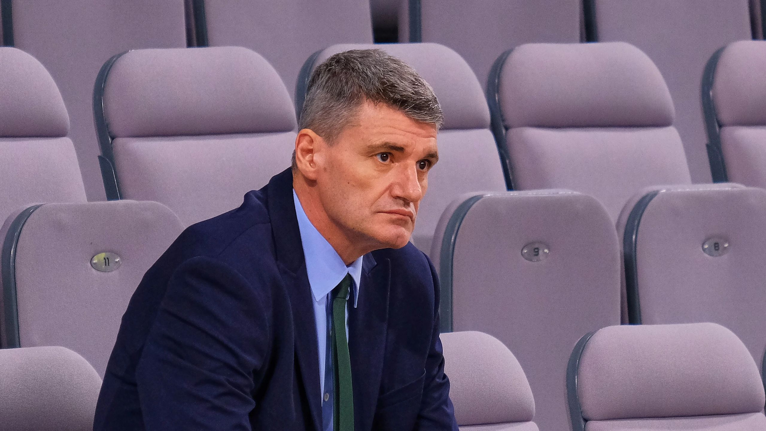 Velimir Perasović, con traje azul y corbata verde, sentado en la grada de un pabellón como entrenador del UNICS Kazán, en marzo de 2025